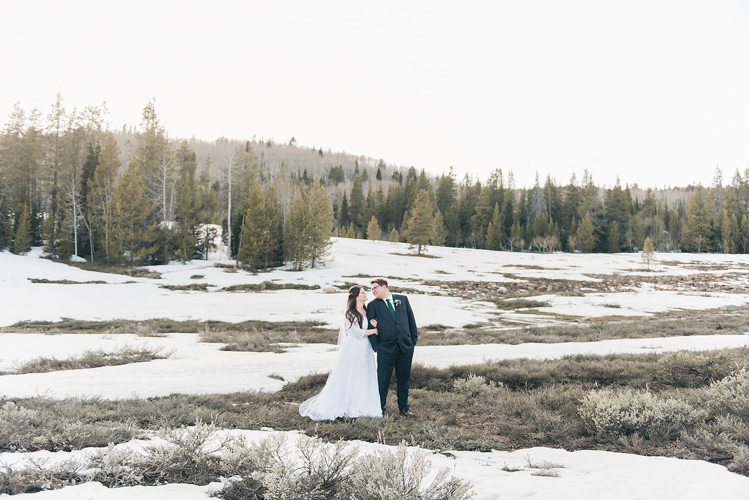 Bride and groom stand together in a snow-covered mountain meadow with forest in background.
