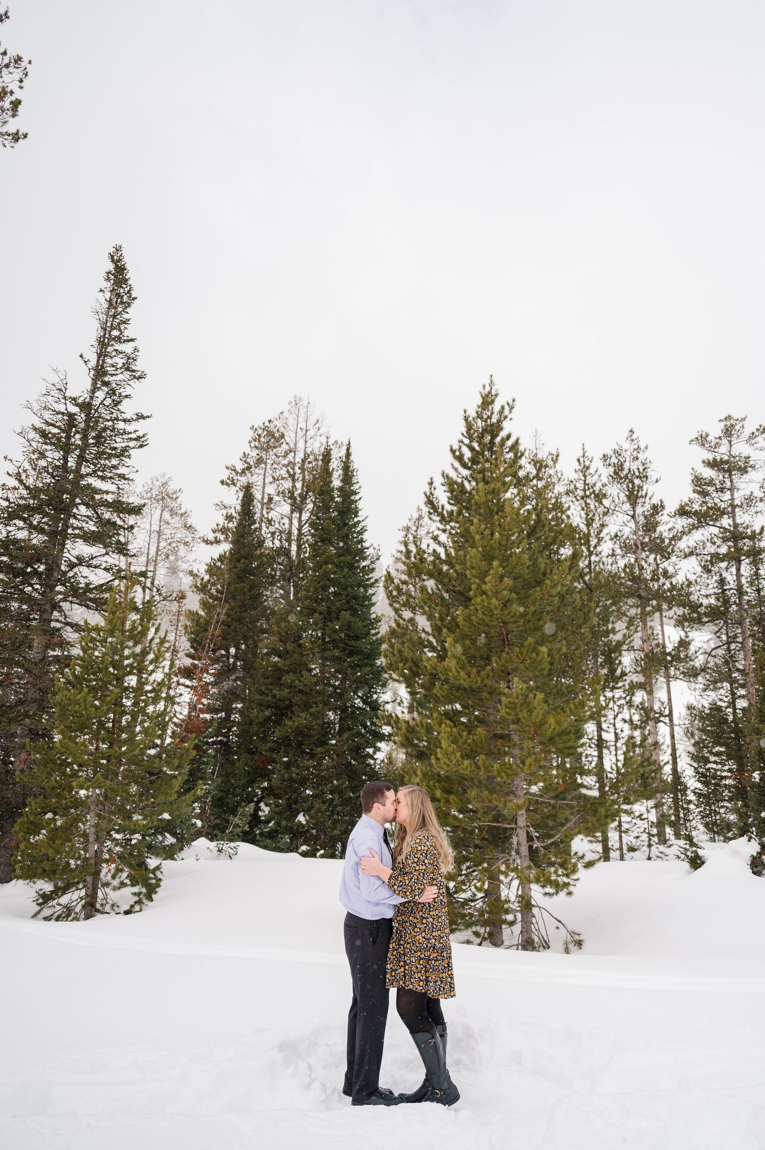 A couple embracing in a snowy winter forest with evergreen trees in the background.