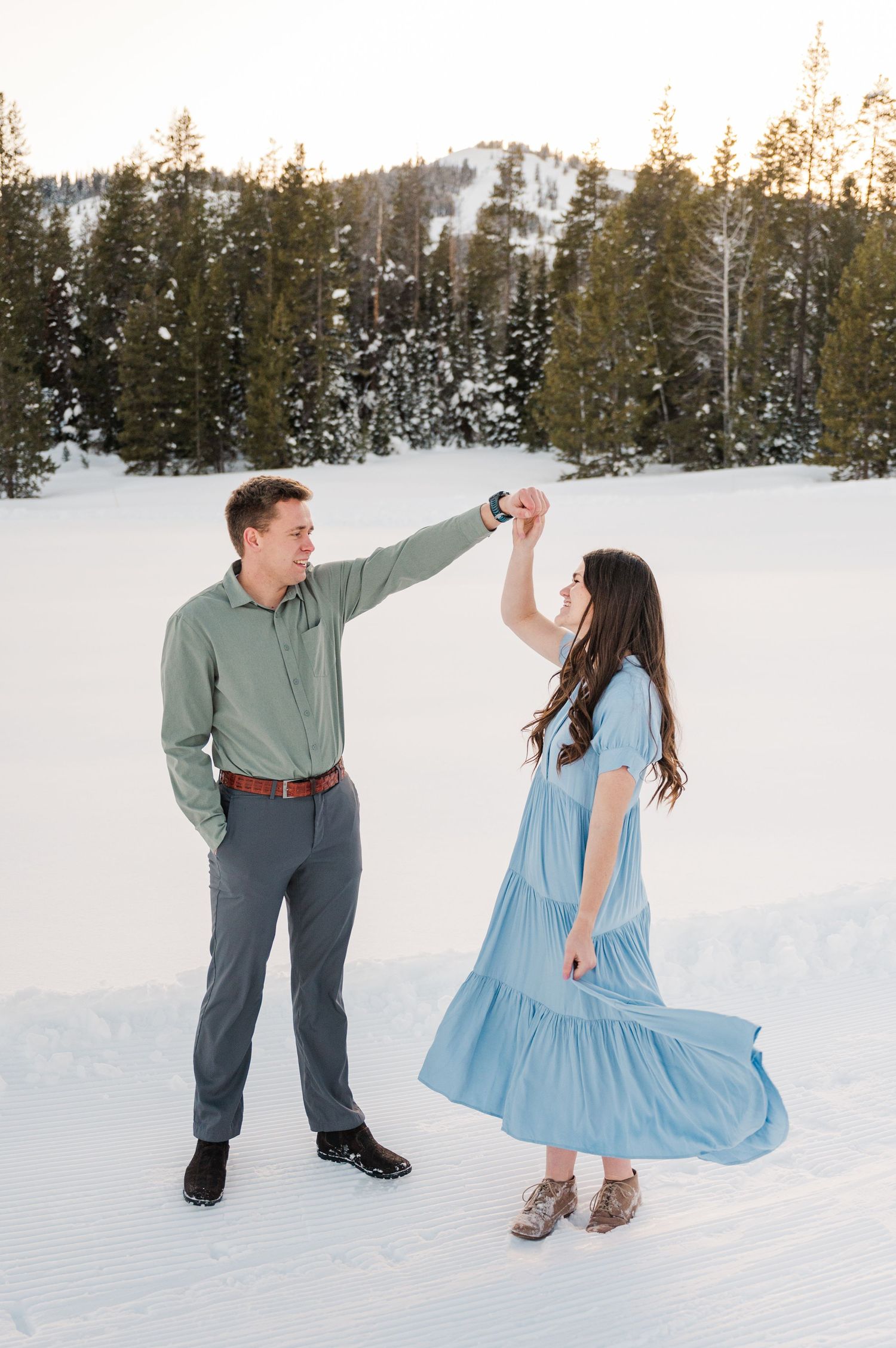 Winter engagement photo of couple dancing in snow with evergreen forest background.