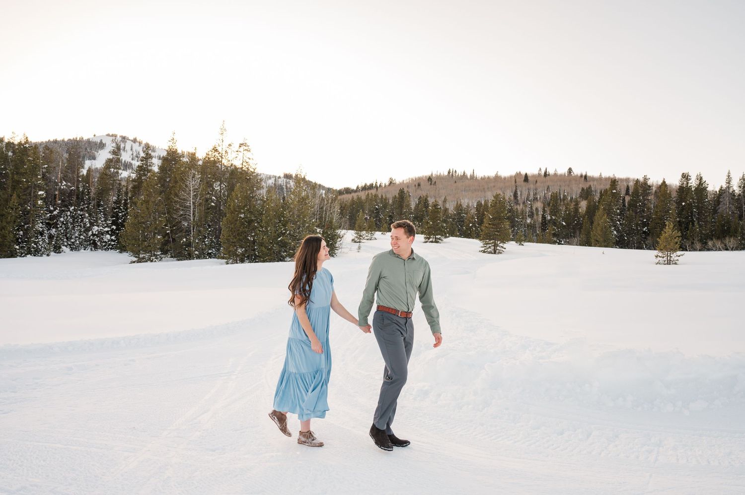 A couple walking hand in hand through a snow-covered field with mountains in the background.
