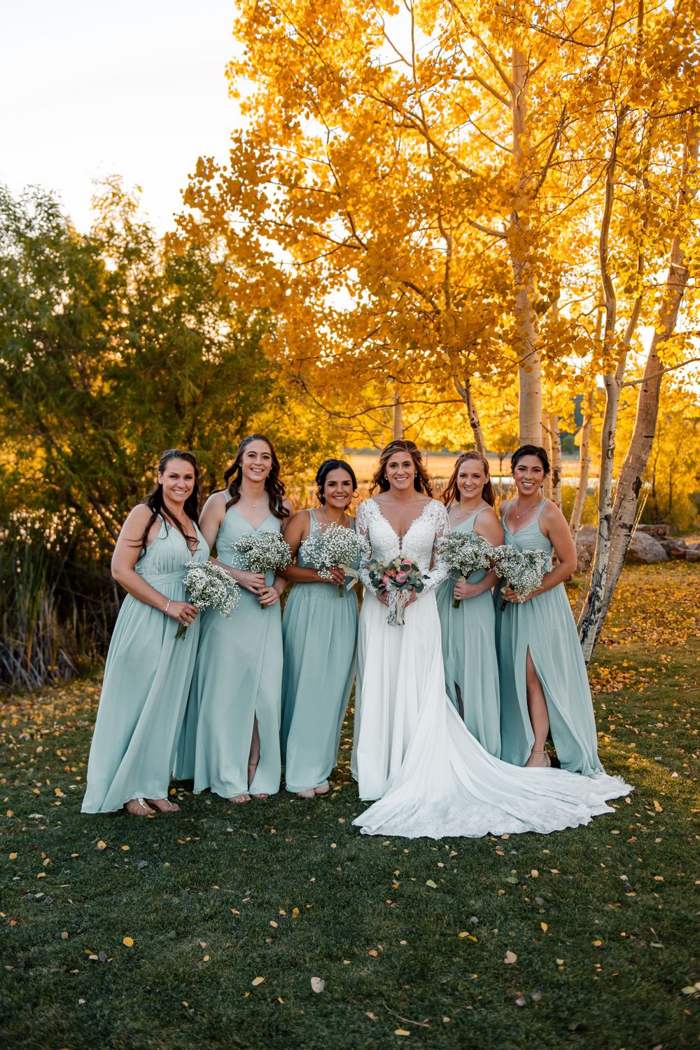 Wedding party in mint green bridesmaid dresses poses against golden autumn trees at sunset.