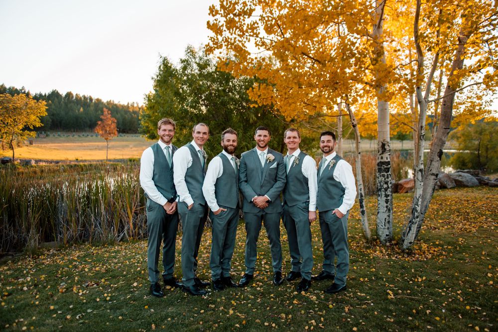Groomsmen in matching gray vests pose together on an autumn lawn with golden trees in the background.