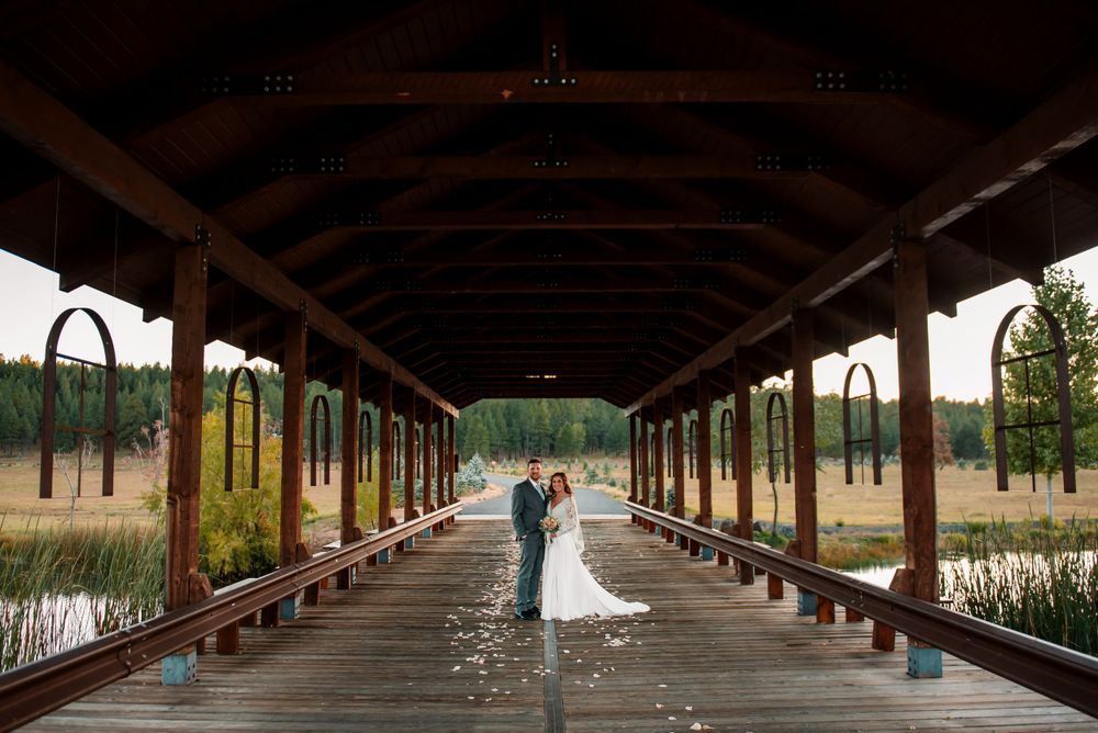 A wedding couple stands at the end of a long wooden covered bridge over water.