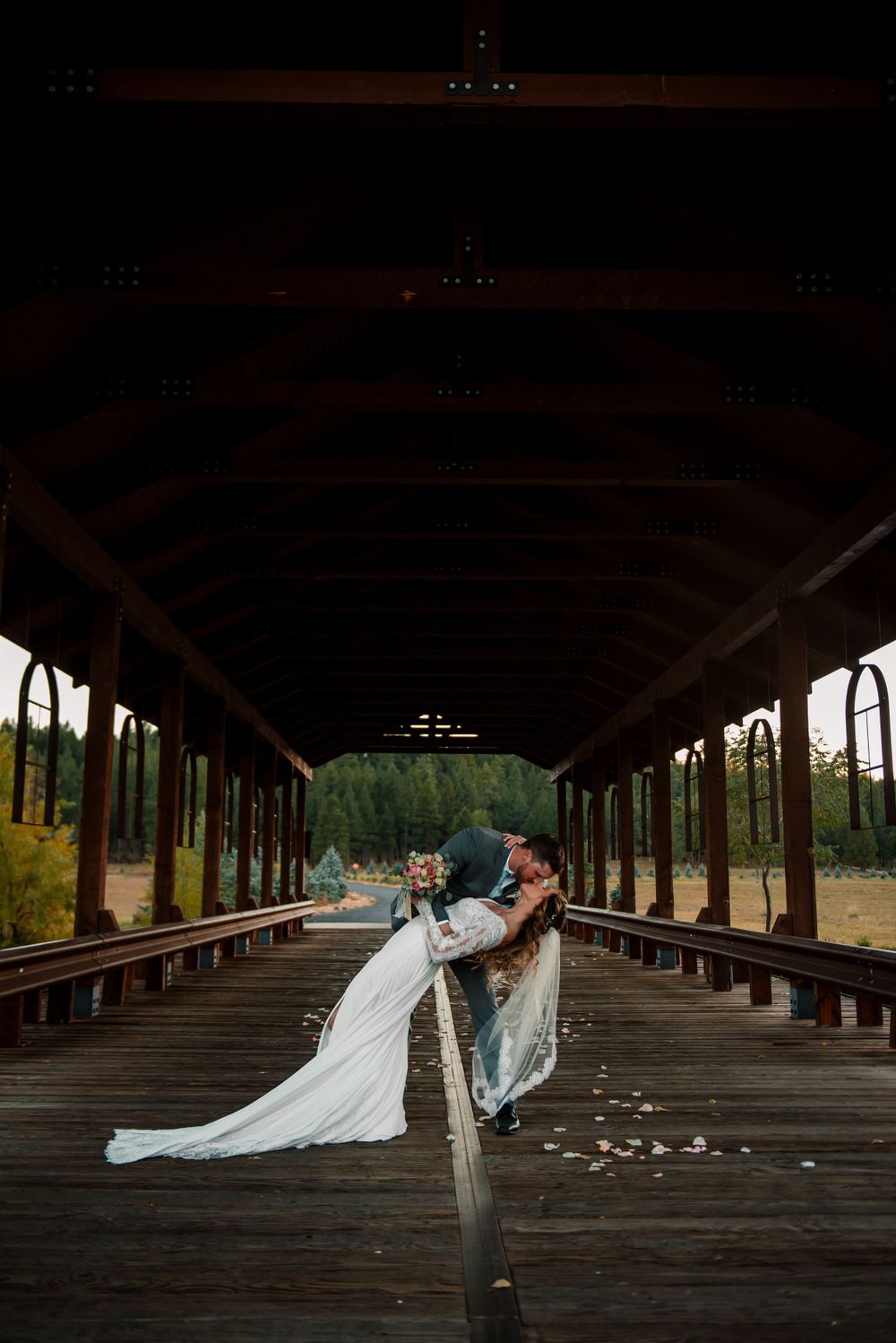 Wedding couple shares intimate moments in a long wooden covered bridge with scattered rose petals.