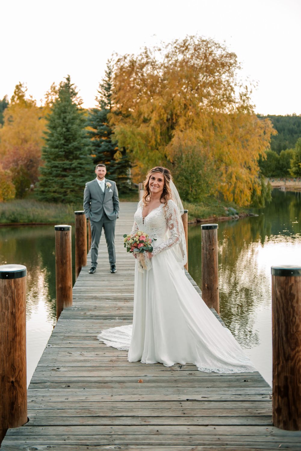 Wedding couple poses on wooden dock with fall foliage and lake reflections in background.