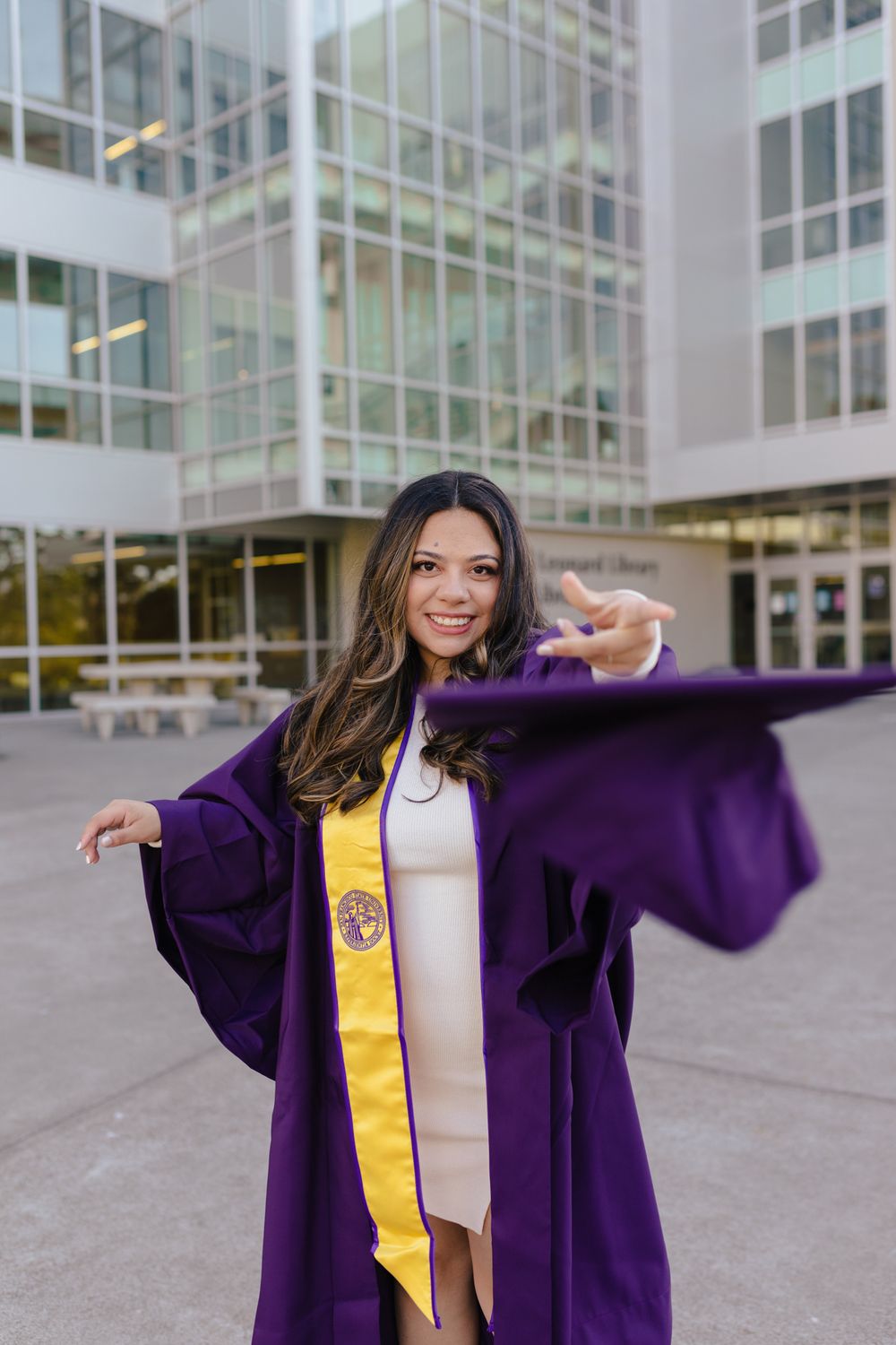 Graduate in purple academic regalia poses joyfully outside modern building.