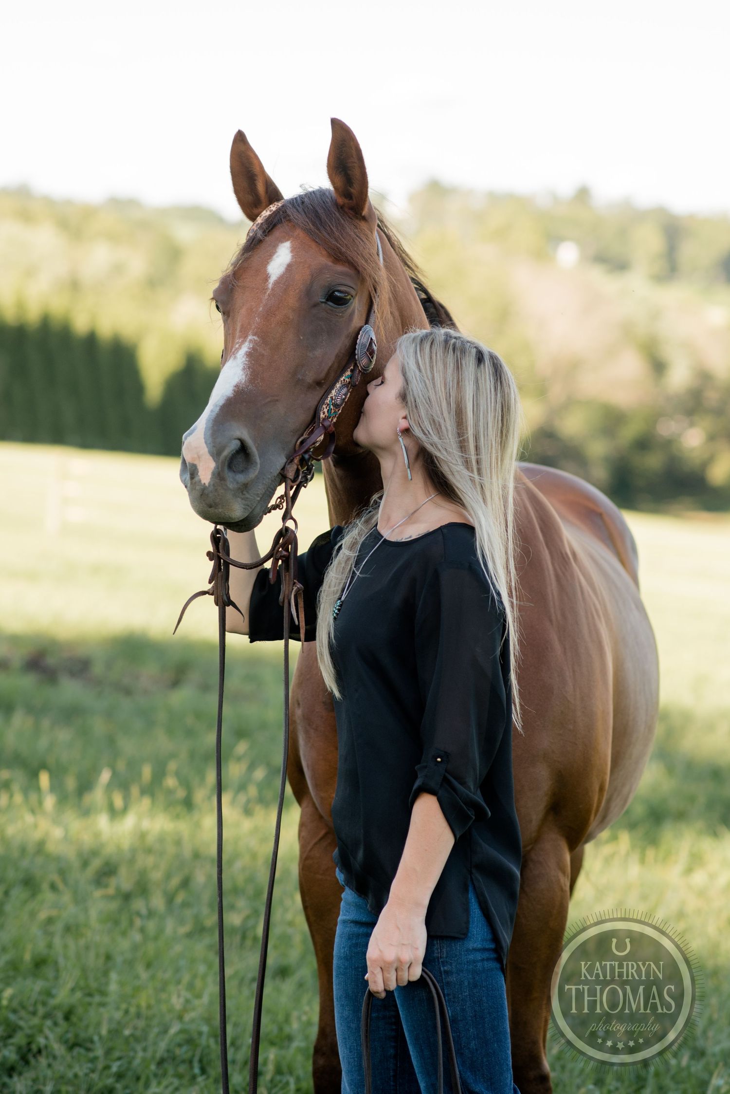 Bay horse with bridle stands close to someone in black top in grassy field.