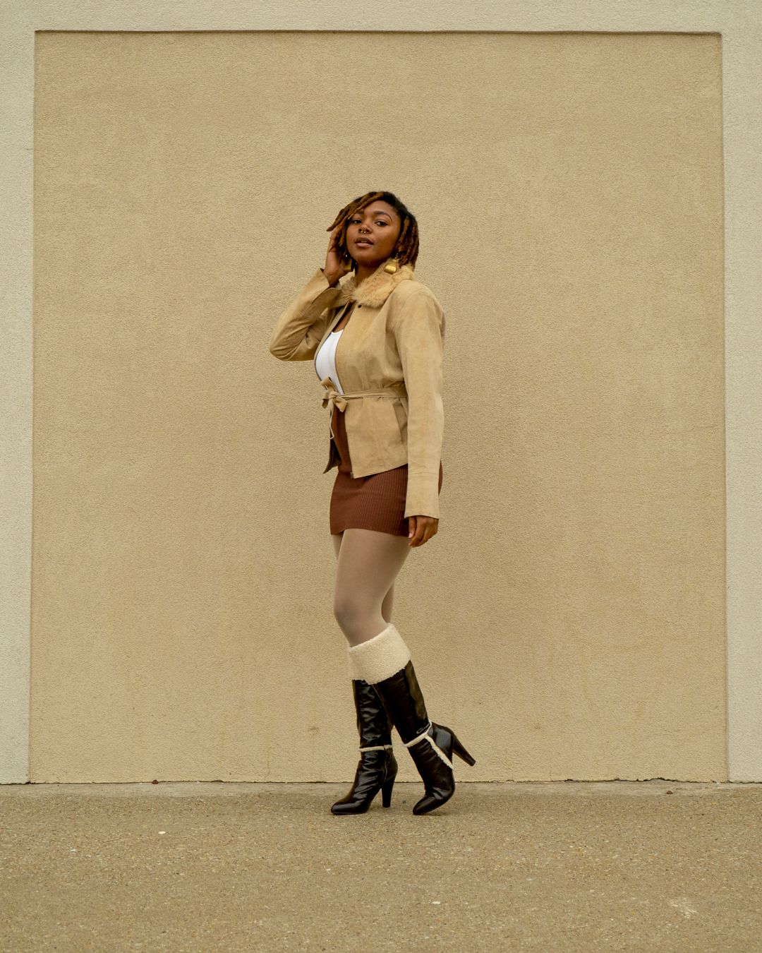 A fashion photograph showing a cream blazer, shorts, and black boots against a beige wall background.