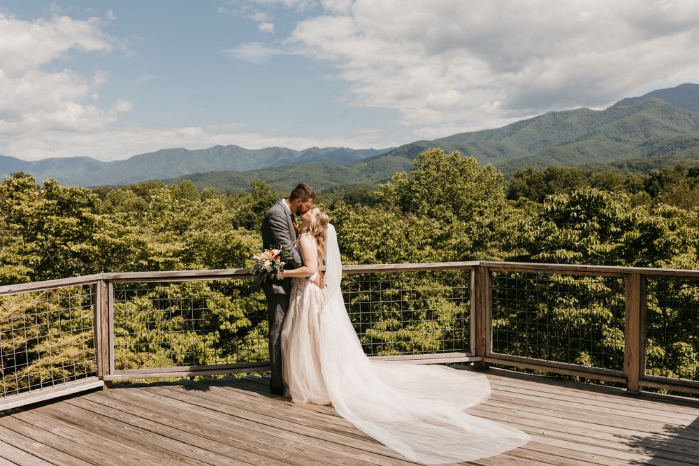 Bride and groom embrace on scenic wooden deck overlooking lush green mountains under cloudy sky.