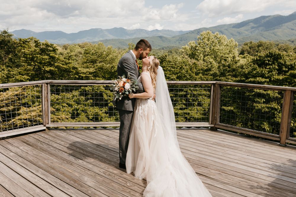 Bride and groom embrace tenderly on a wooden deck with stunning mountain landscape in the background.
