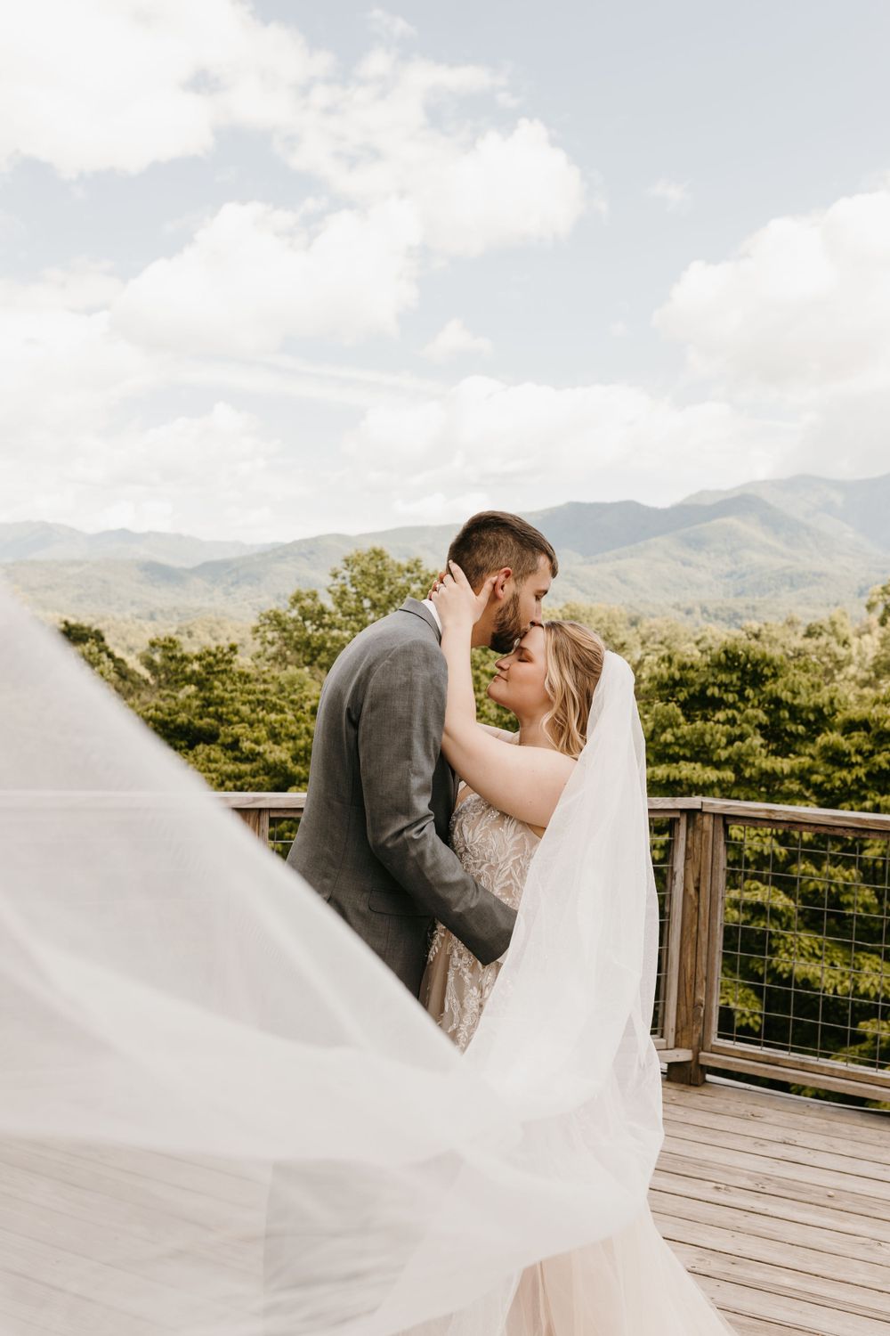Bride and groom lean in for a kiss as her long veil billows in the breeze with mountains behind them.