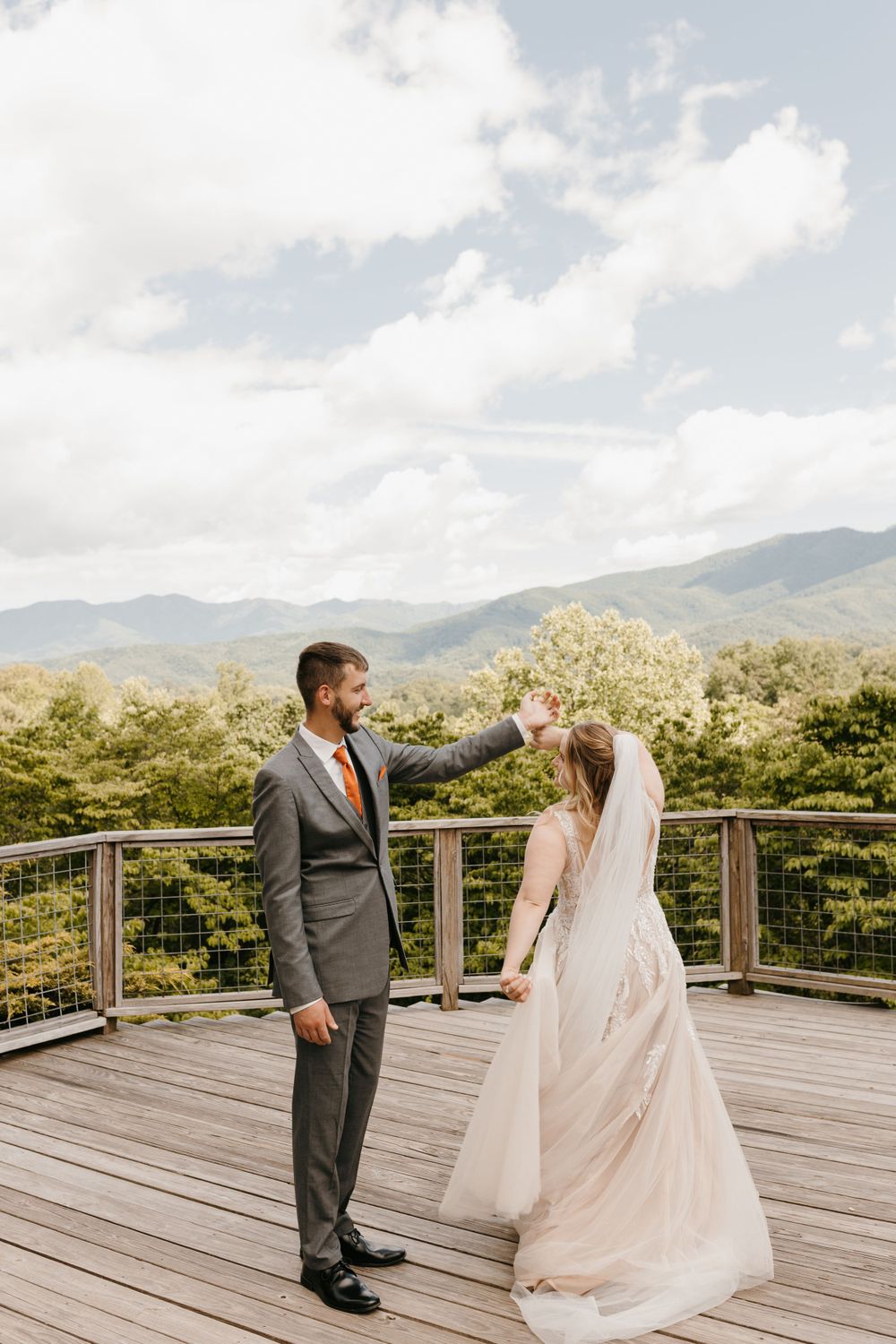 Groom spins bride under his arm on a sunlit deck with lush green mountains in the background.