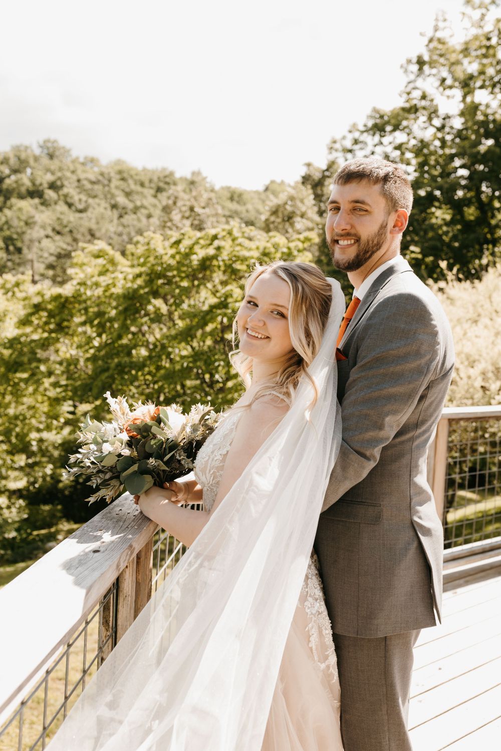 Smiling bride holding a bouquet stands with groom on a scenic outdoor deck surrounded by green trees.