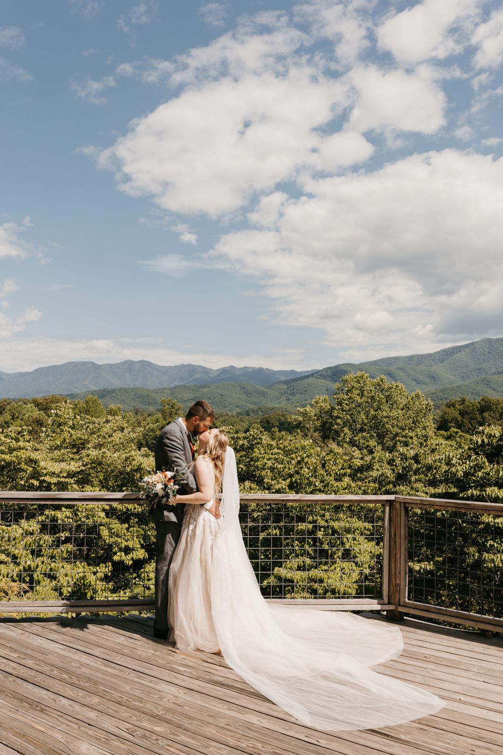 Bride and groom share a kiss on a wooden deck overlooking lush mountain views on their wedding day.