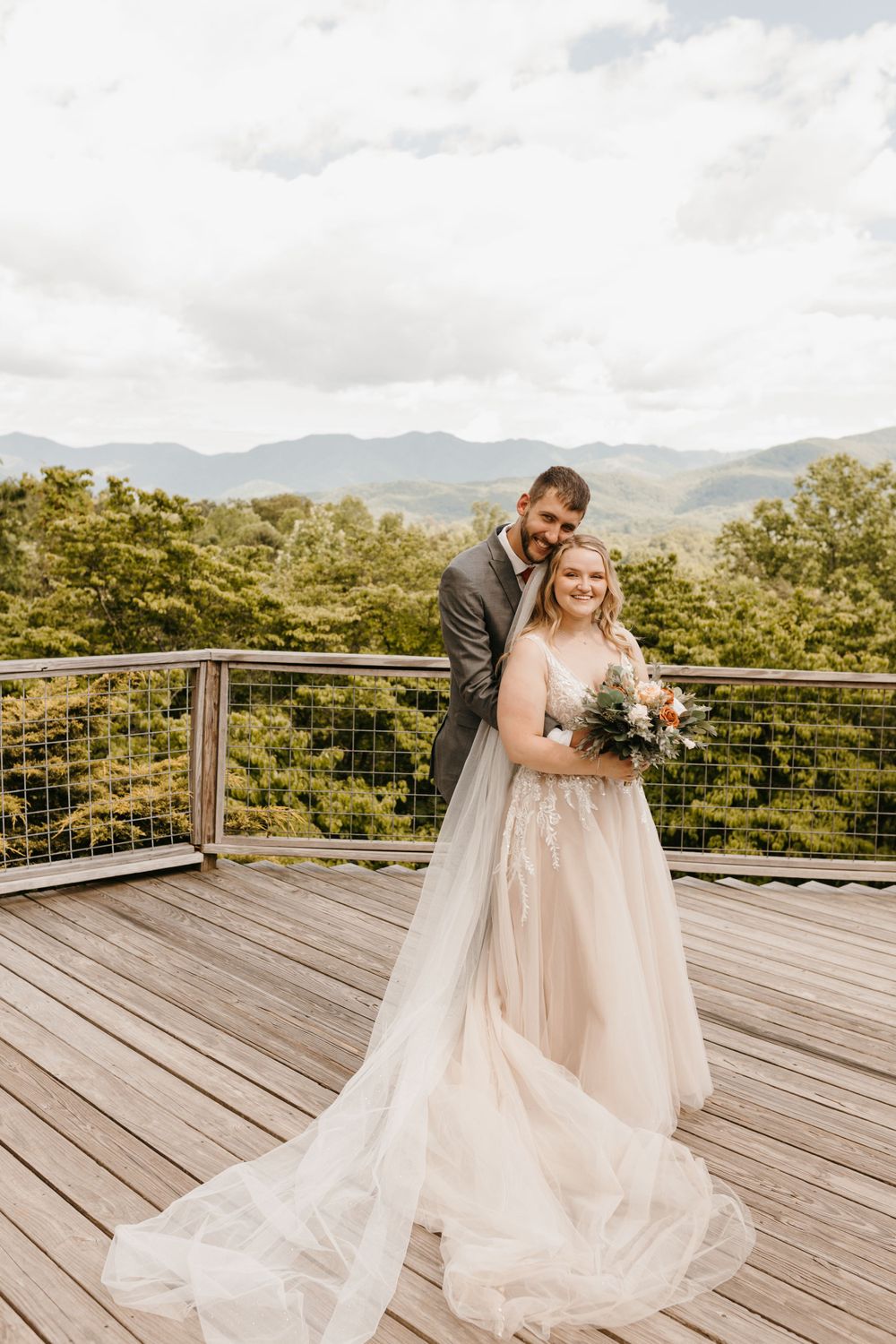 Bride and groom pose together on a wooden deck, her long veil trailing as mountains frame the scene.