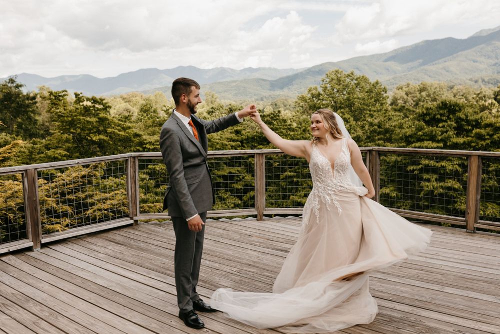 Groom twirls bride in her flowing wedding gown on a wooden deck with scenic mountain views.
