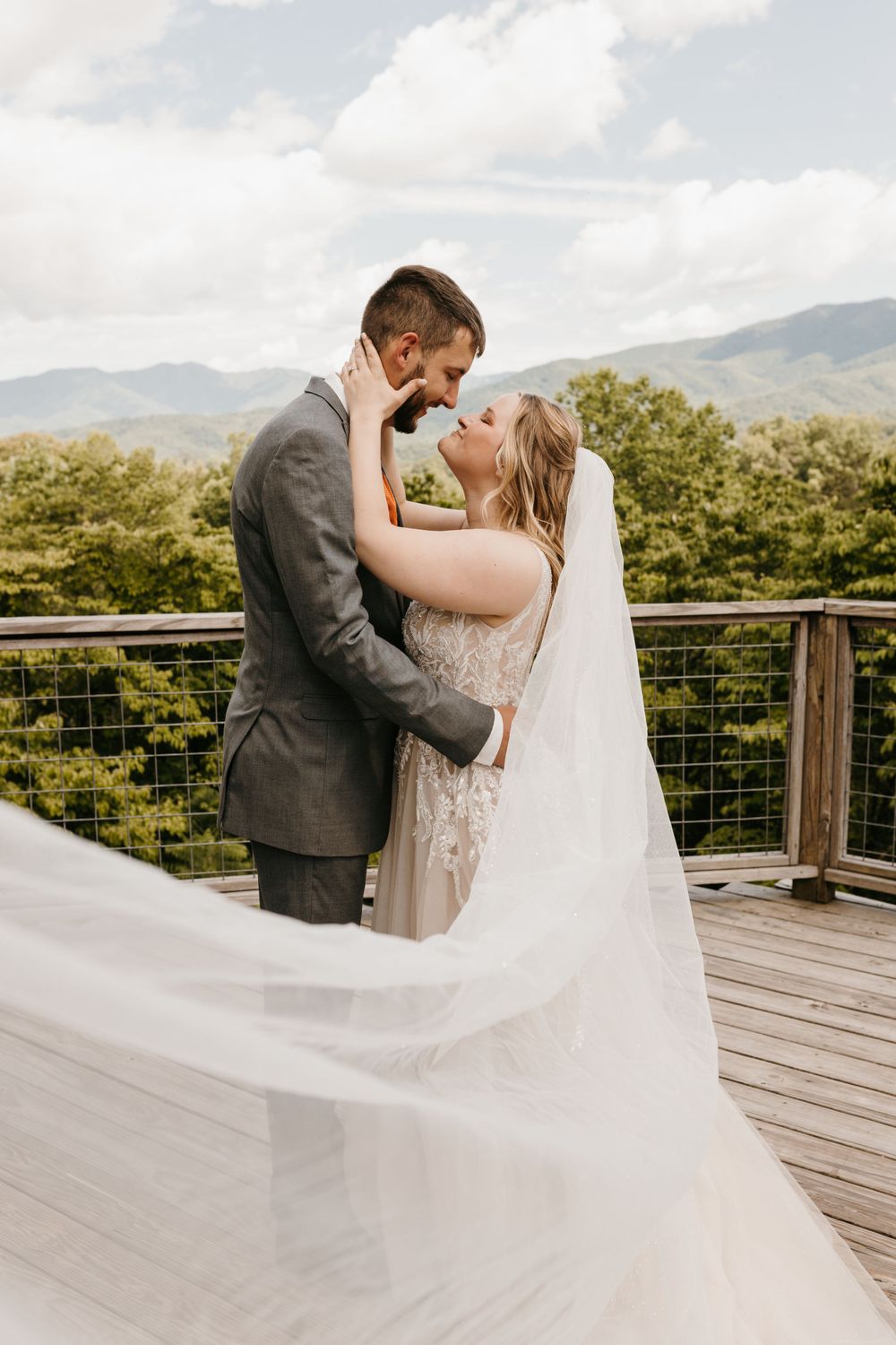 Bride and groom share a romantic moment as her veil flows in the wind against a mountain backdrop.