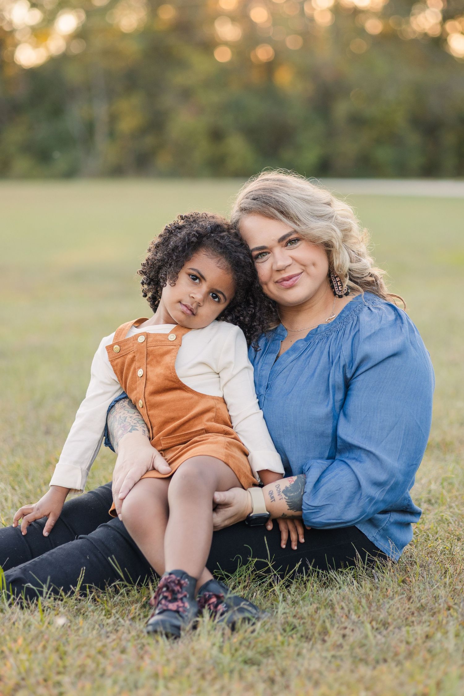 mother in a blue shirt holds daughter in field in an orange outfit during golden hour