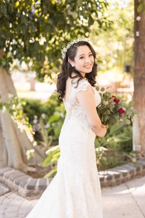 Bride in lace dress poses with bouquet in garden setting with flowering trees.
