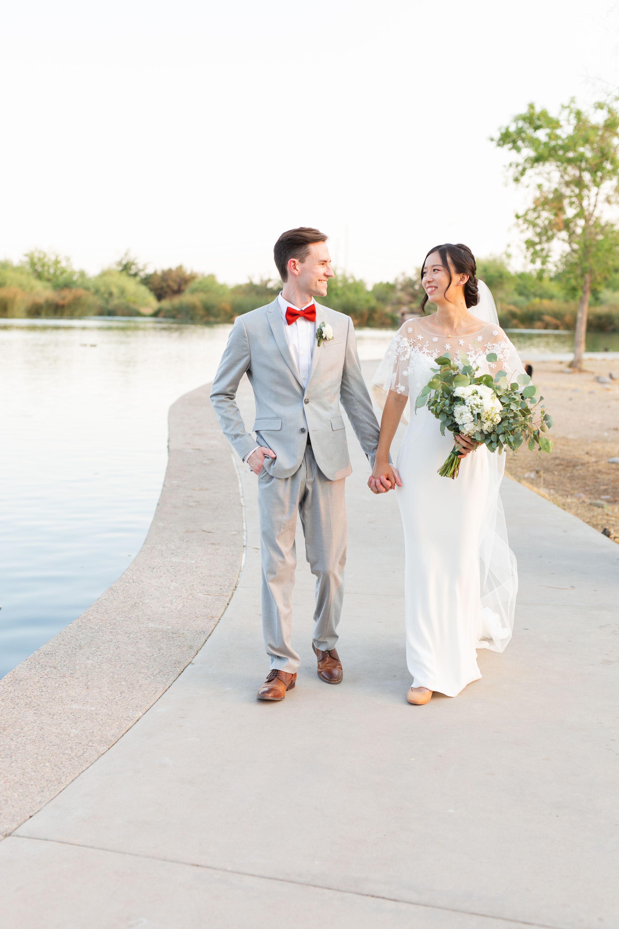 Newlyweds walk hand in hand along waterfront path, groom in light grey suit with red bowtie.