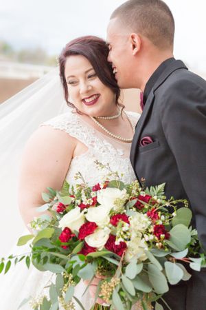 Elegant wedding couple embracing outdoors with a stunning red and white floral bouquet.