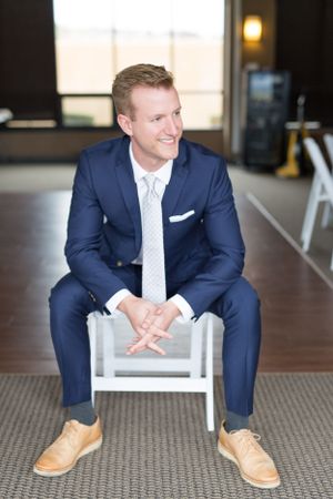 Groom in navy suit sits casually on white chair before ceremony.