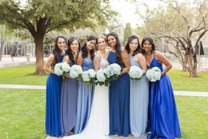 Bridal party in coordinated blue dresses holds matching hydrangea bouquets on grassy lawn.