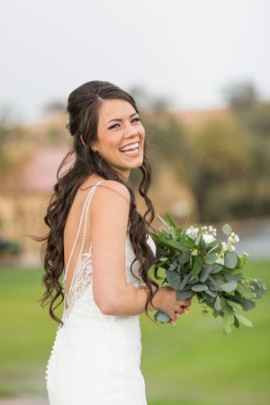 Bride with long dark curls holds green and white bouquet while laughing outdoors.