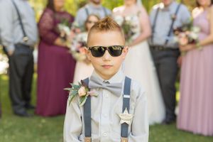 Young ring bearer in suspenders and bow tie wearing sunglasses at outdoor wedding.