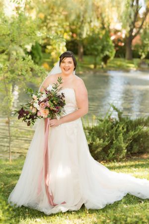 Bride in flowing white gown holds gorgeous bouquet with pink ribbons by garden pond.