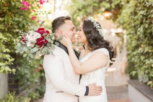 Romantic wedding couple embraces in garden pathway surrounded by pink bougainvillea flowers.
