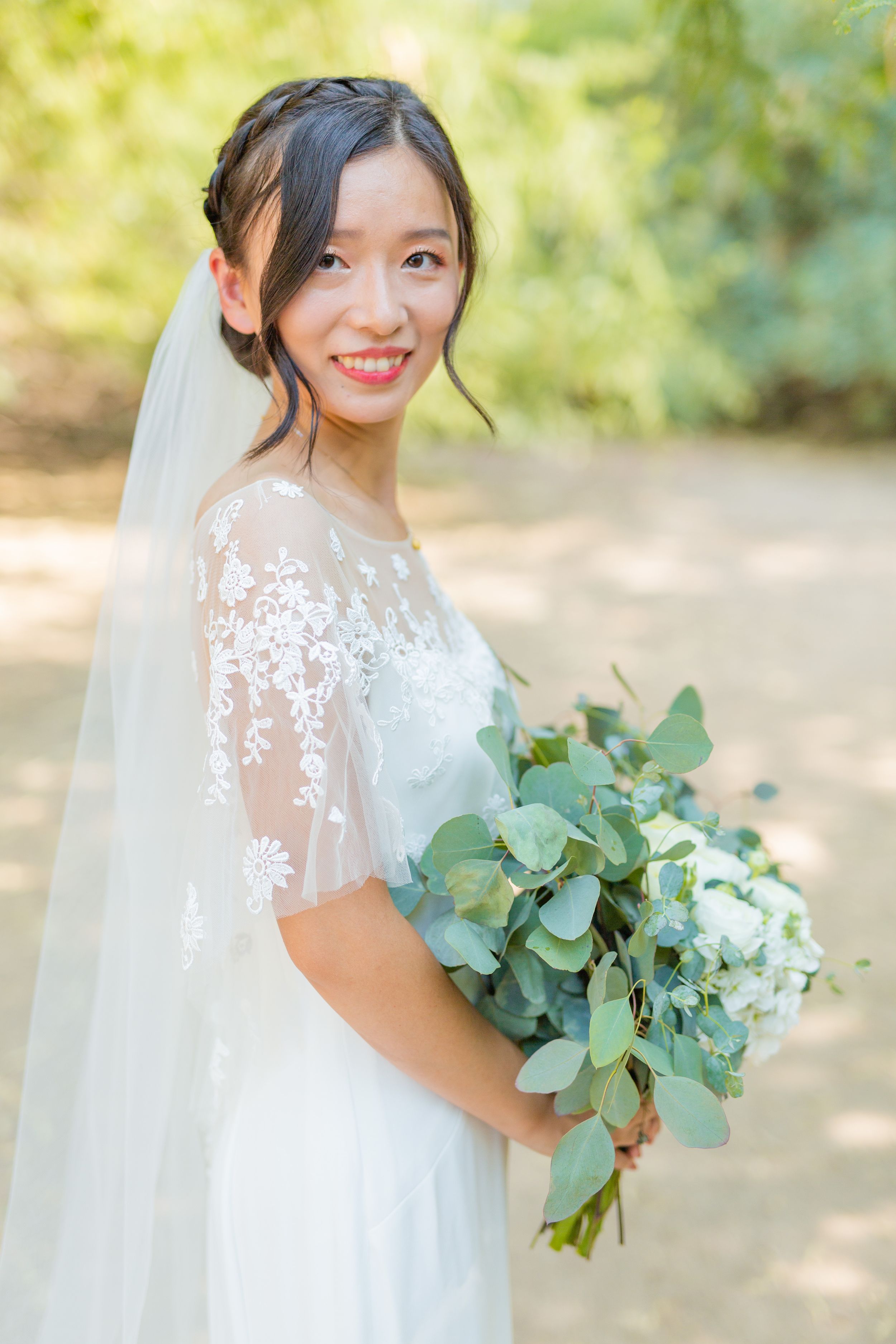 Bride in lace gown holds eucalyptus bouquet against sunlit natural backdrop.