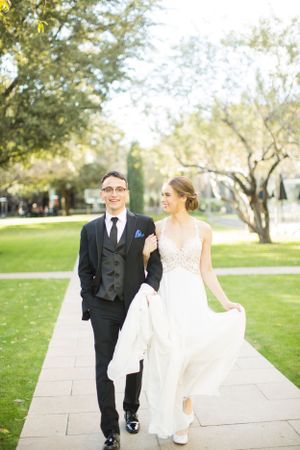 Elegant couple walks along stone pathway surrounded by green lawn and trees.