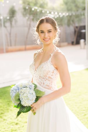 Bride in lace wedding dress holds blue hydrangea bouquet in sunlit garden setting.