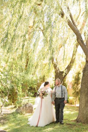 Wedding couple poses under romantic weeping willow tree in garden setting.