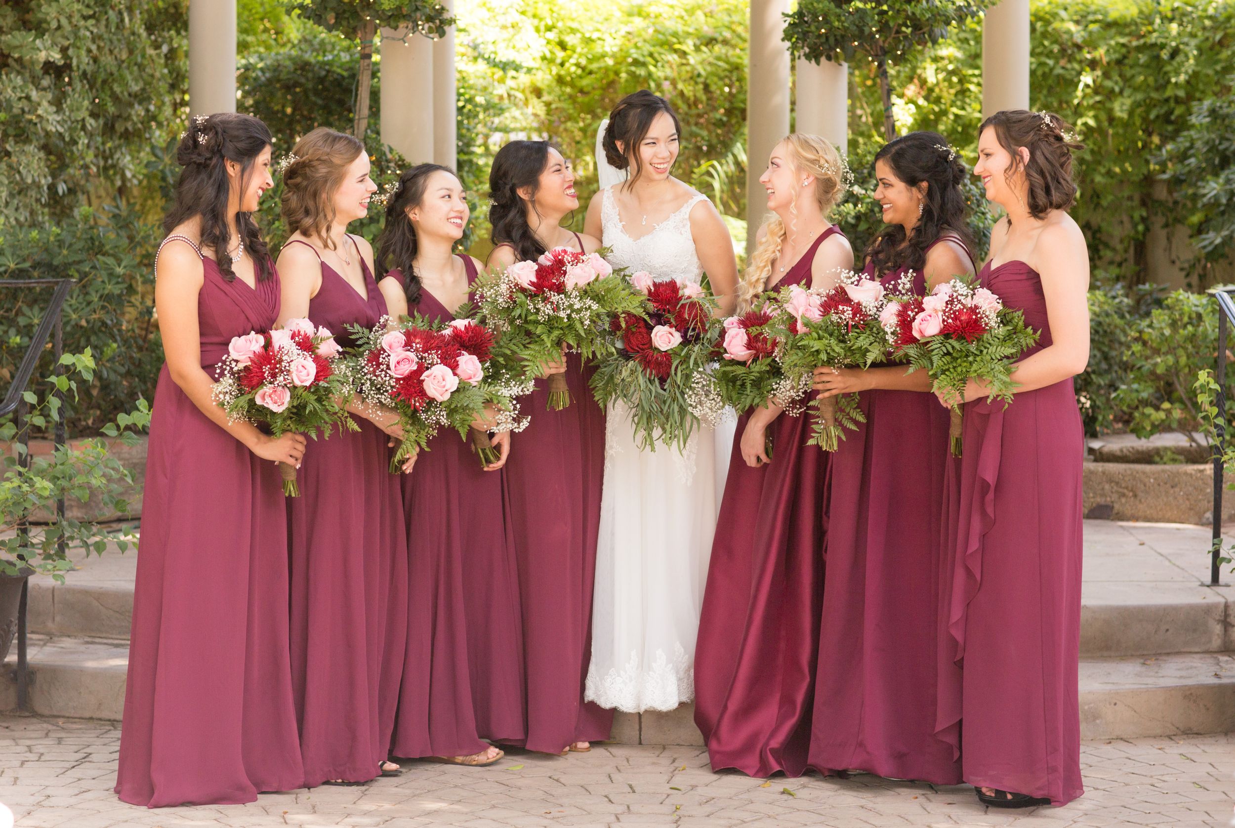 Bridesmaids in burgundy dresses hold pink and red bouquets while posing under columns.