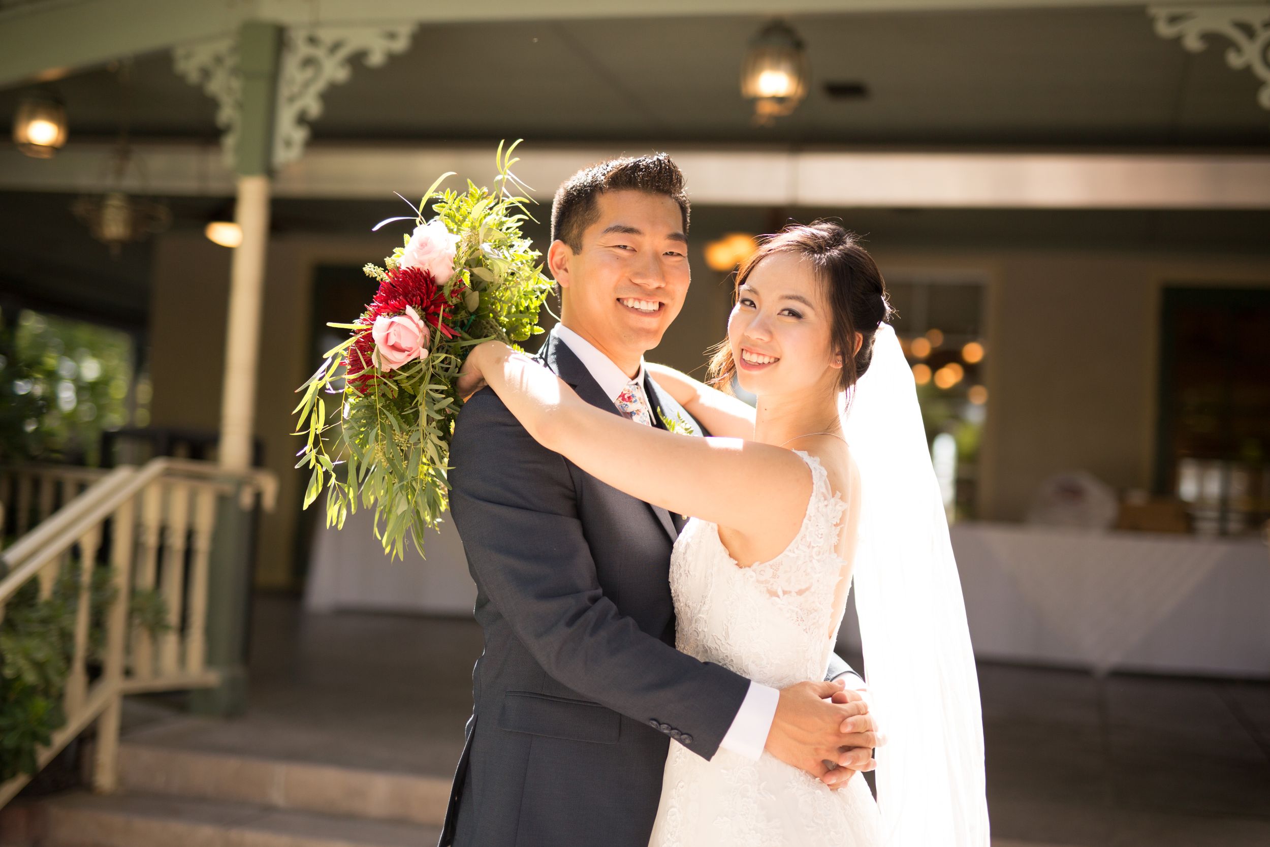 Wedding couple embraces joyfully while holding vibrant red and white bouquet under decorative pavilion.