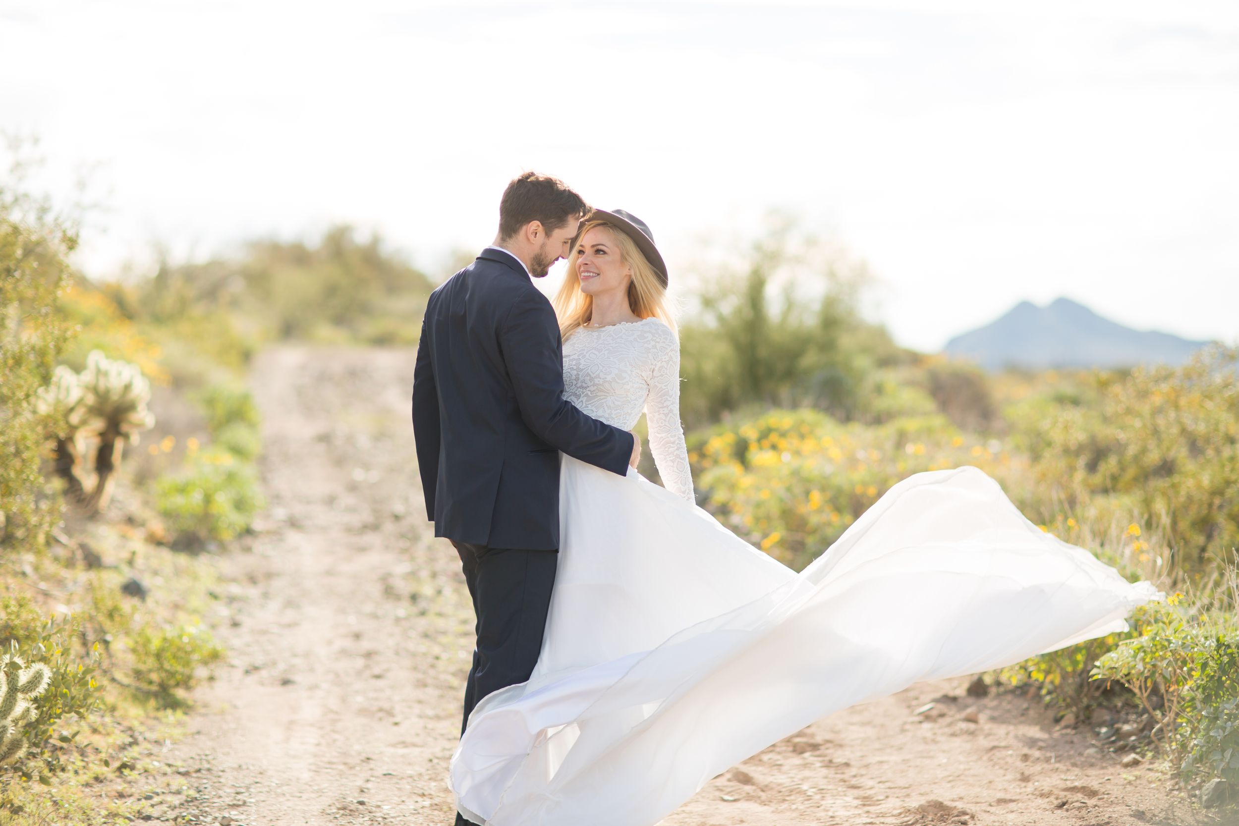 A couple in wedding attire embraces on a desert trail with mountains in the background.