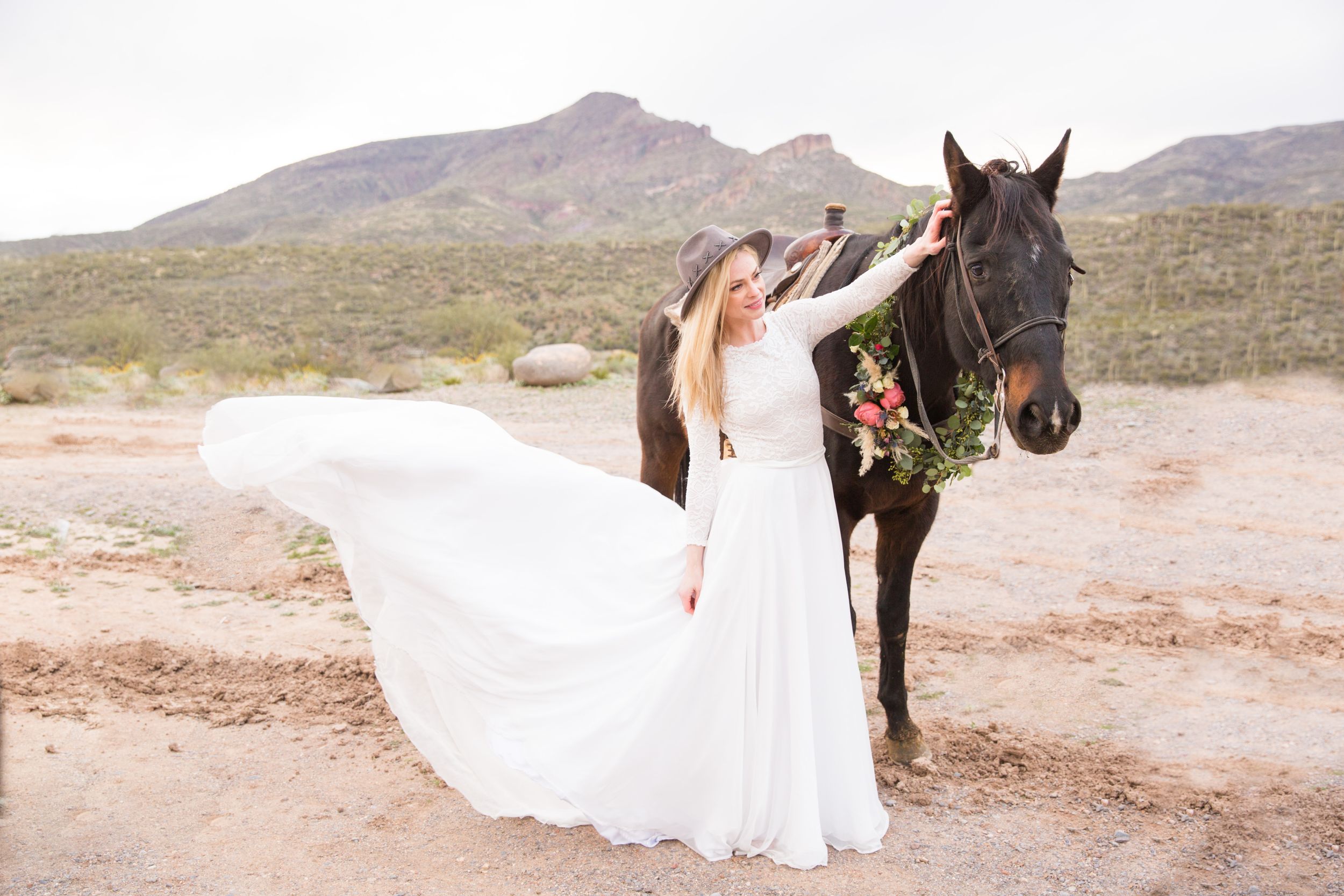 A flowing white wedding dress billows in the wind next to a decorated horse in a desert landscape.