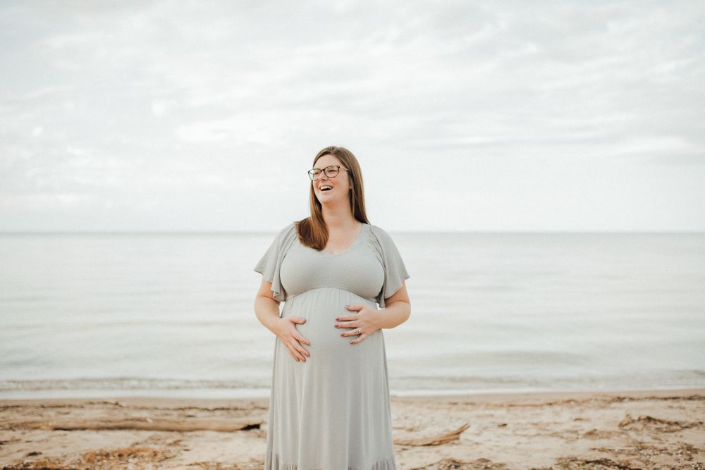 A joyful maternity moment captured on a cloudy beach in a flowing gray dress.