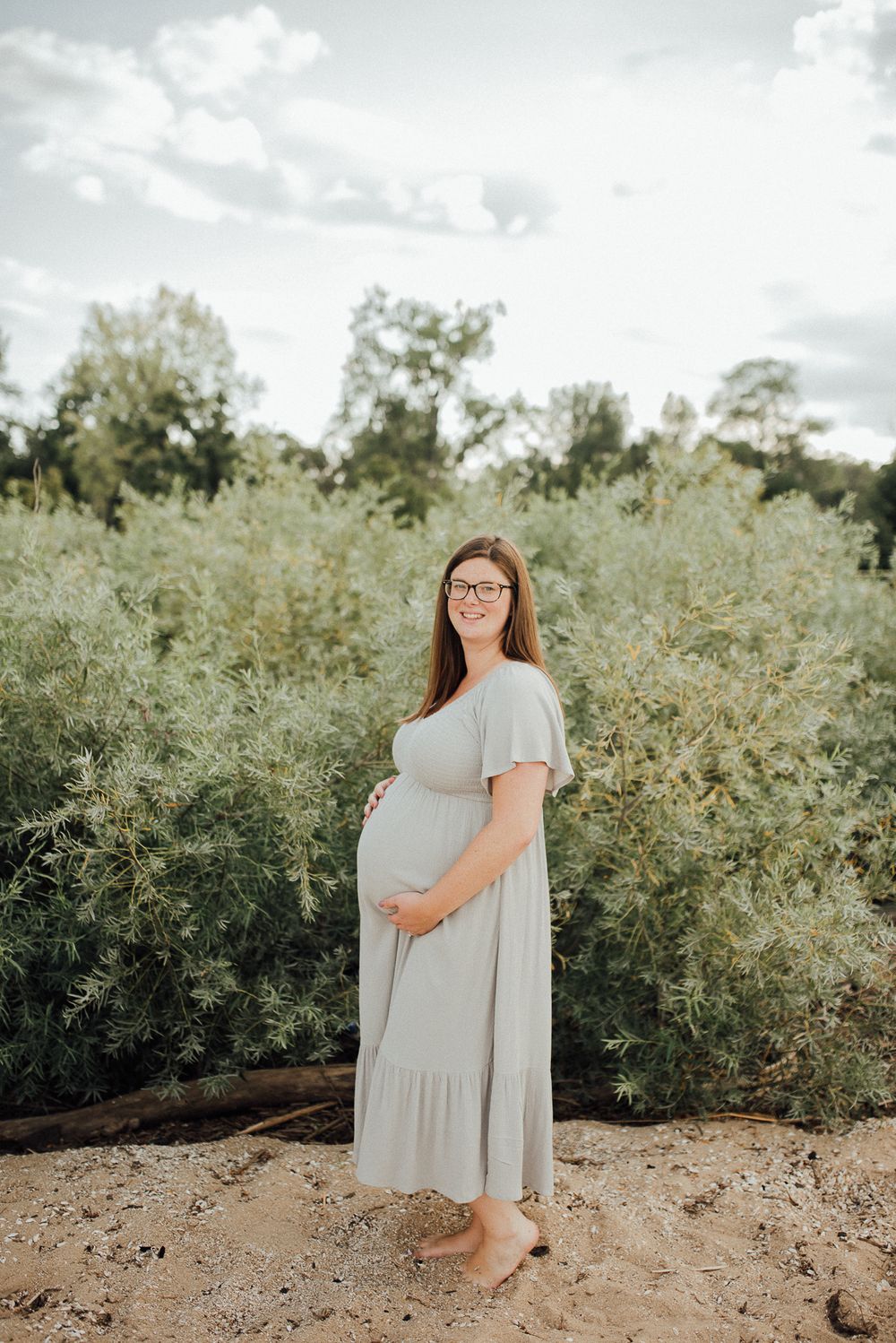 A maternity portrait in a light gray dress standing barefoot against greenery and cloudy skies.