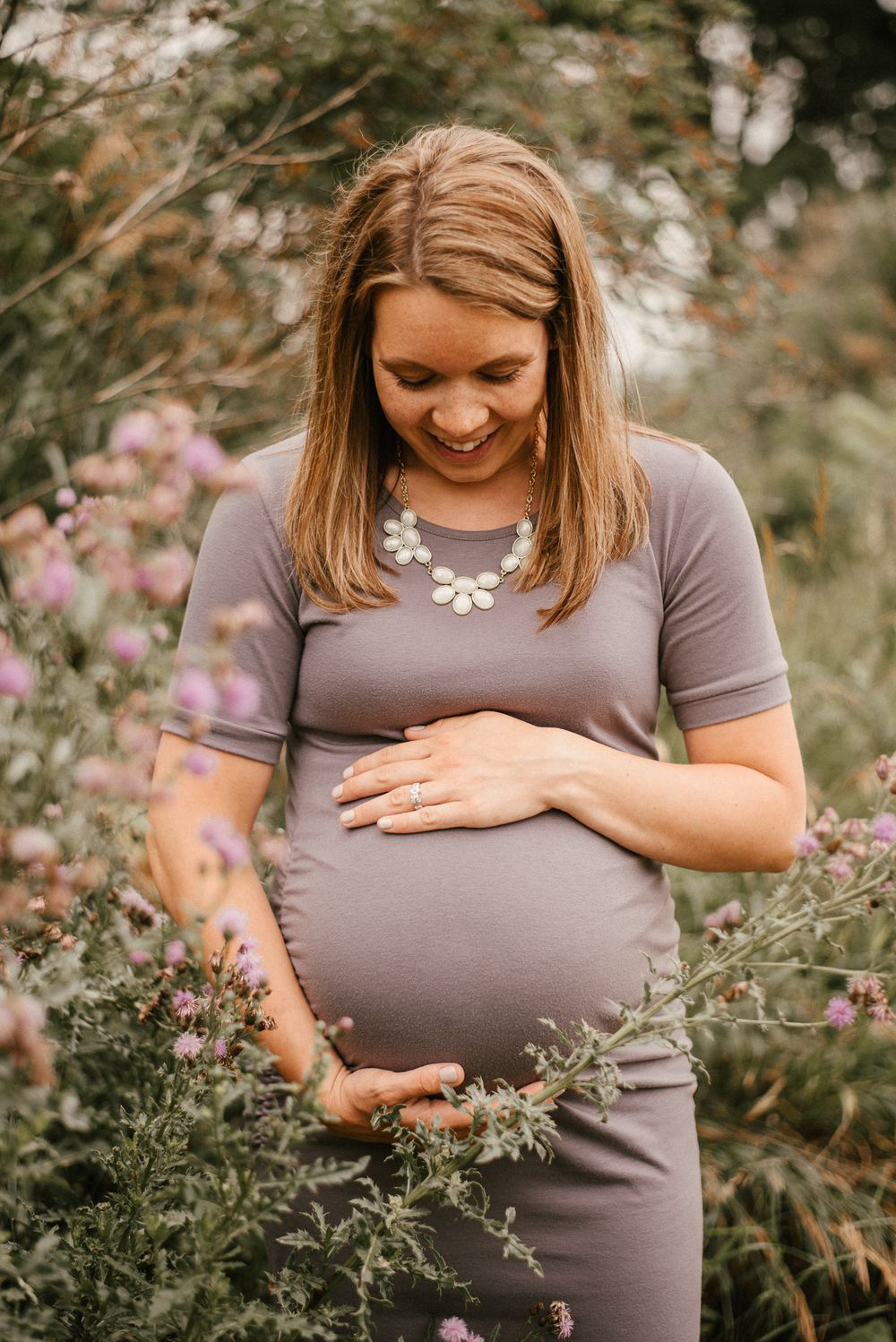 Expectant mother in purple dress smiling while standing among wildflowers.