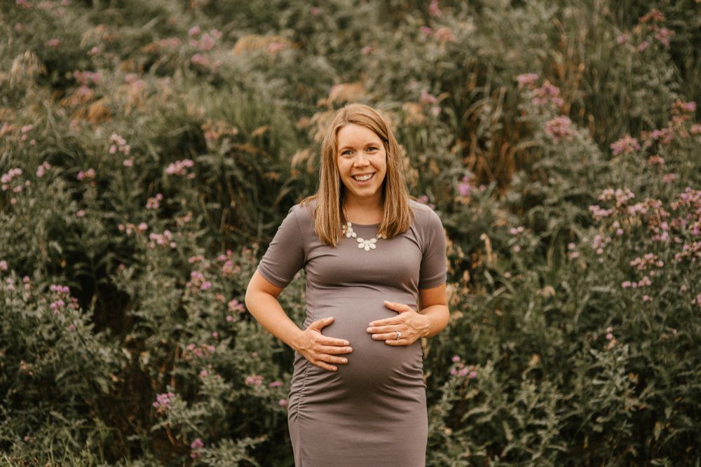 A maternity photo showing a gray dress against a background of lush green foliage and pink flowers.