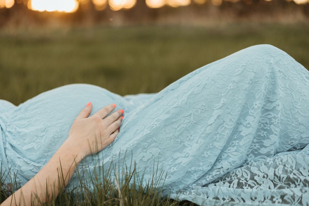 A close-up of a baby bump draped in ethereal pale blue lace against sunset lighting.