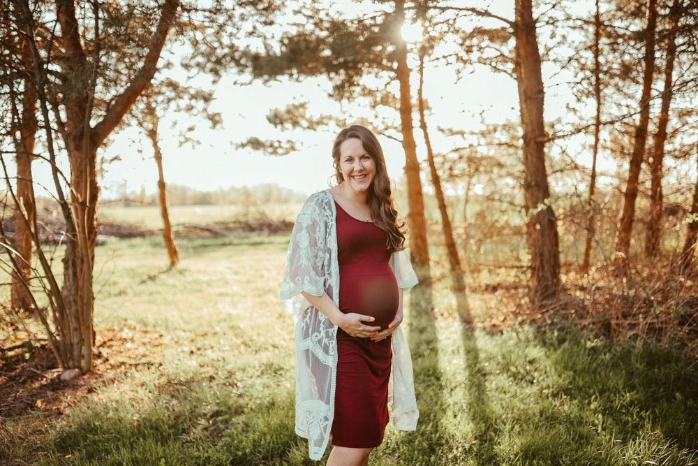 A maternity portrait in a burgundy dress with sunlight filtering through autumn trees.