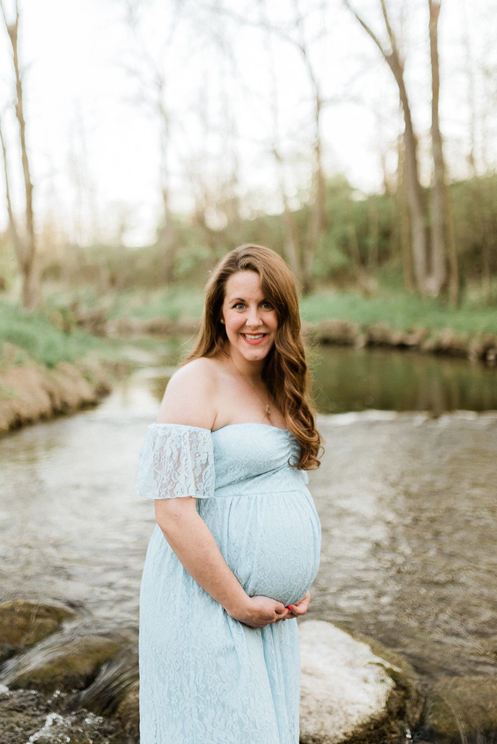A maternity portrait in a pale blue off-shoulder dress by a serene creek.