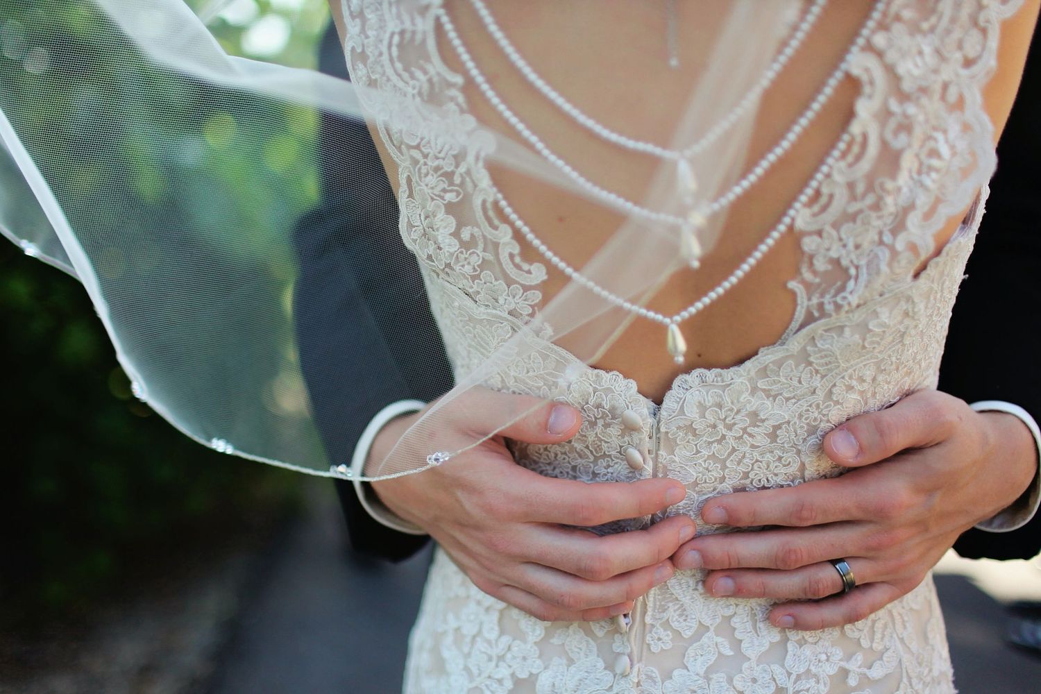 Close-up of bride's hands adjusting lace back of wedding dress with sheer veil visible.