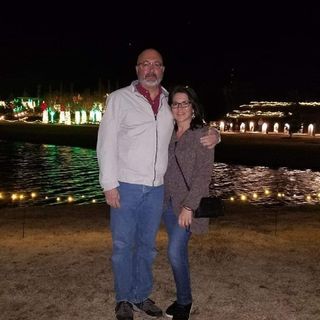 Holiday scene at night with two people posing in front of luminous Christmas light displays near water.