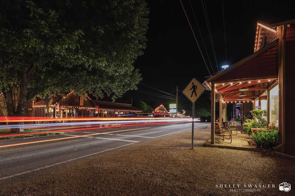 Night street scene with light trails and illuminated storefront in small town setting.