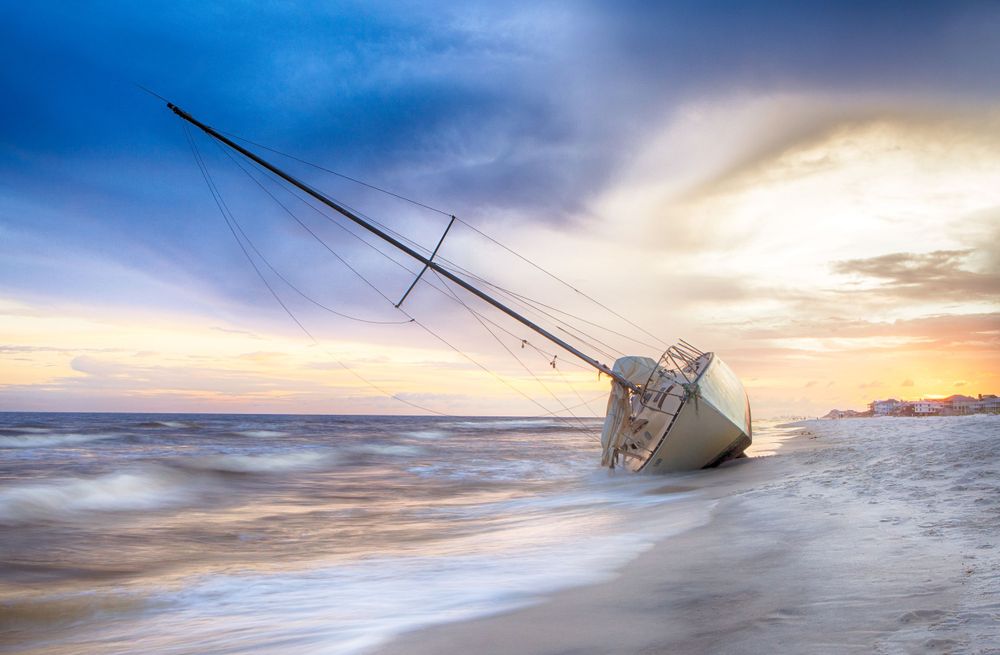 Shipwrecked sailboat partially submerged in ocean waves during a dramatic sunset.